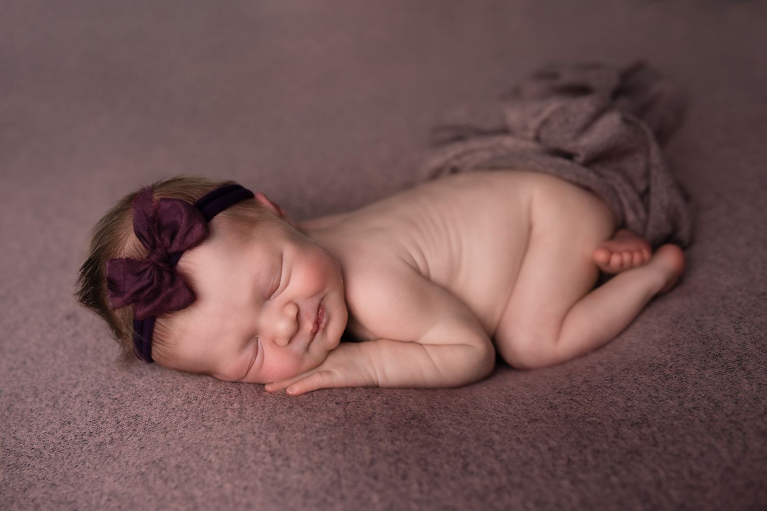 Peaceful newborn baby girl with purple floral headband sleeping on mauve blanket in posed photography session.