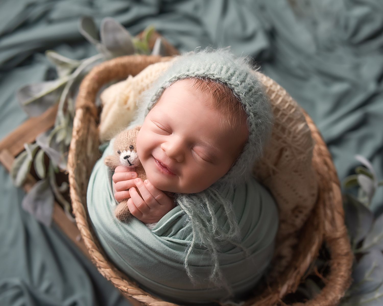 Smiling newborn baby wrapped in teal swaddle holding tiny bear, sleeping in wicker basket with soft green decor.