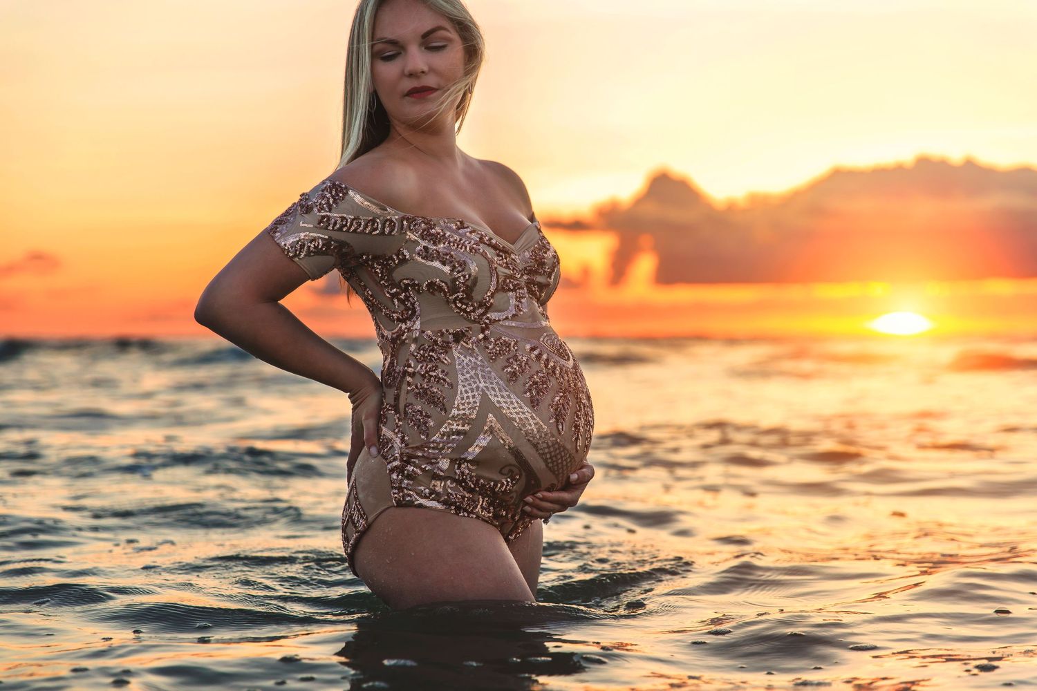 A silhouetted maternity portrait of an expectant mother standing in water during a golden sunset.