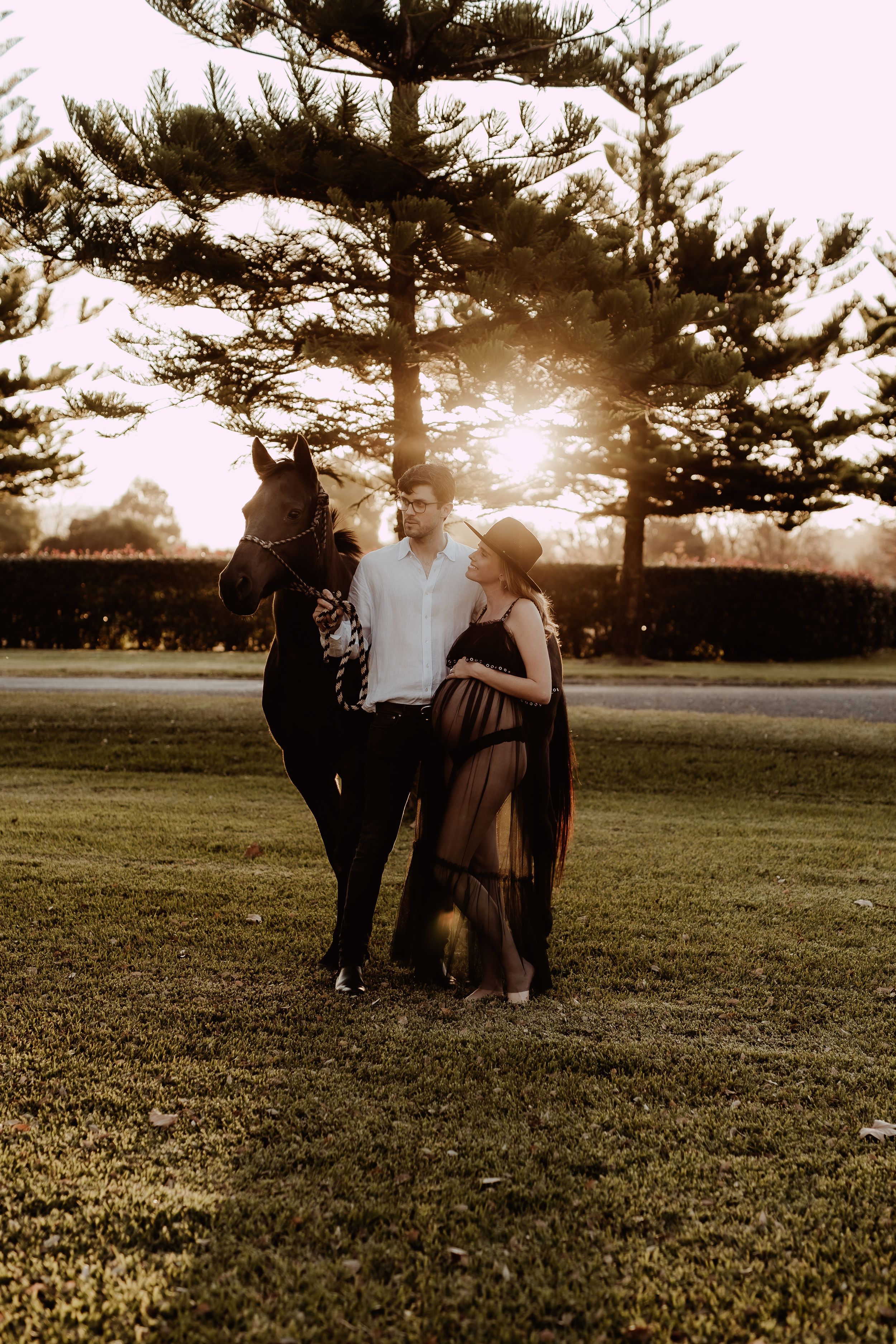 A couple stands together with their dog in a grassy field at sunset with tall pine trees in the background.