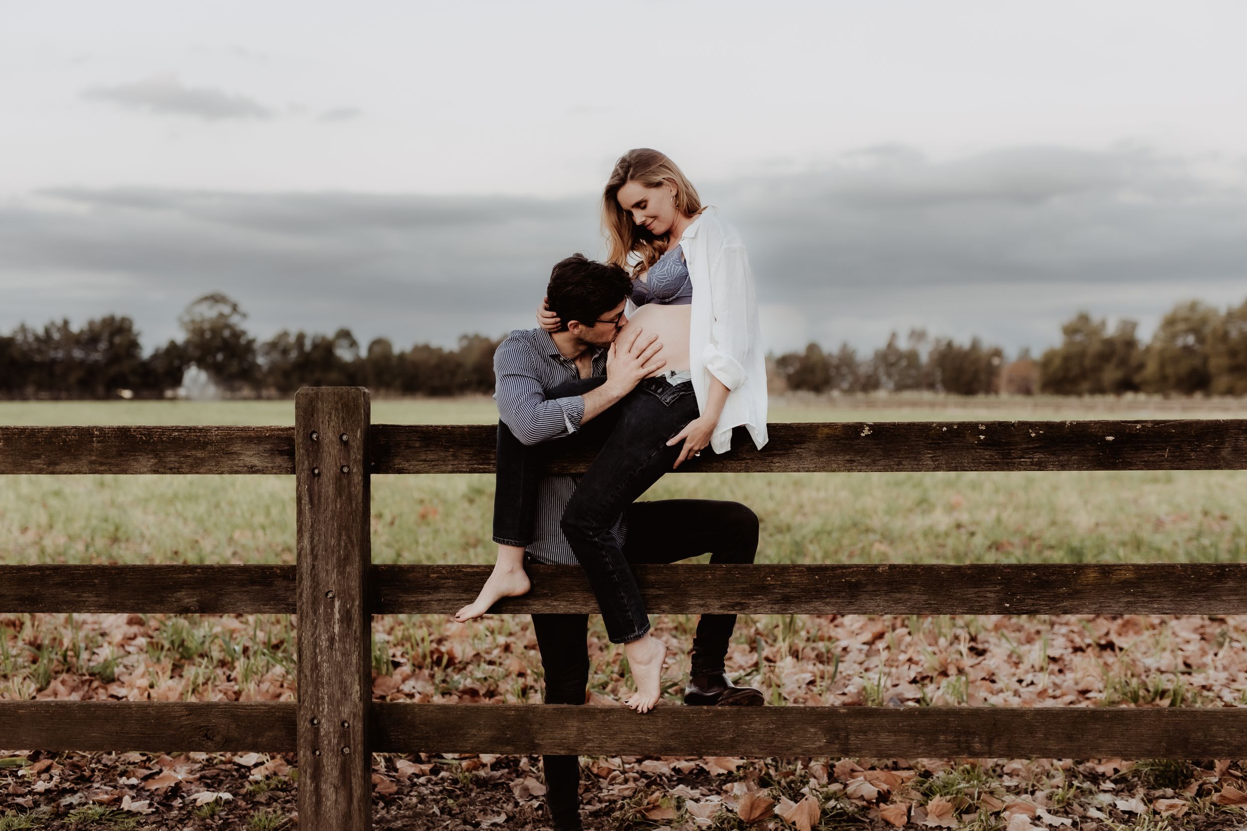 A couple sits together on a wooden fence in a rustic countryside setting.