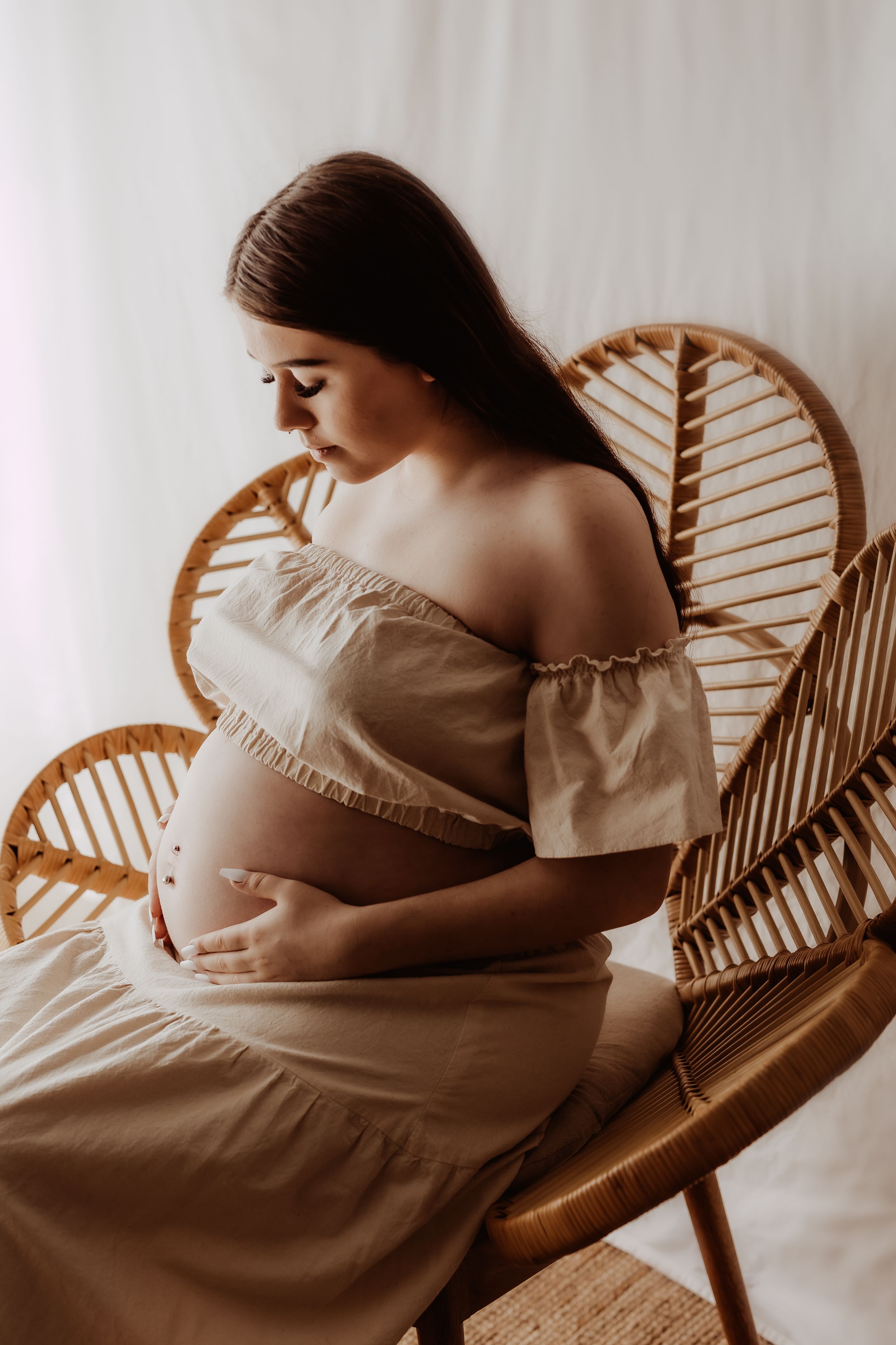 Maternity photoshoot featuring an off-shoulder dress and rattan chair in soft, warm lighting.
