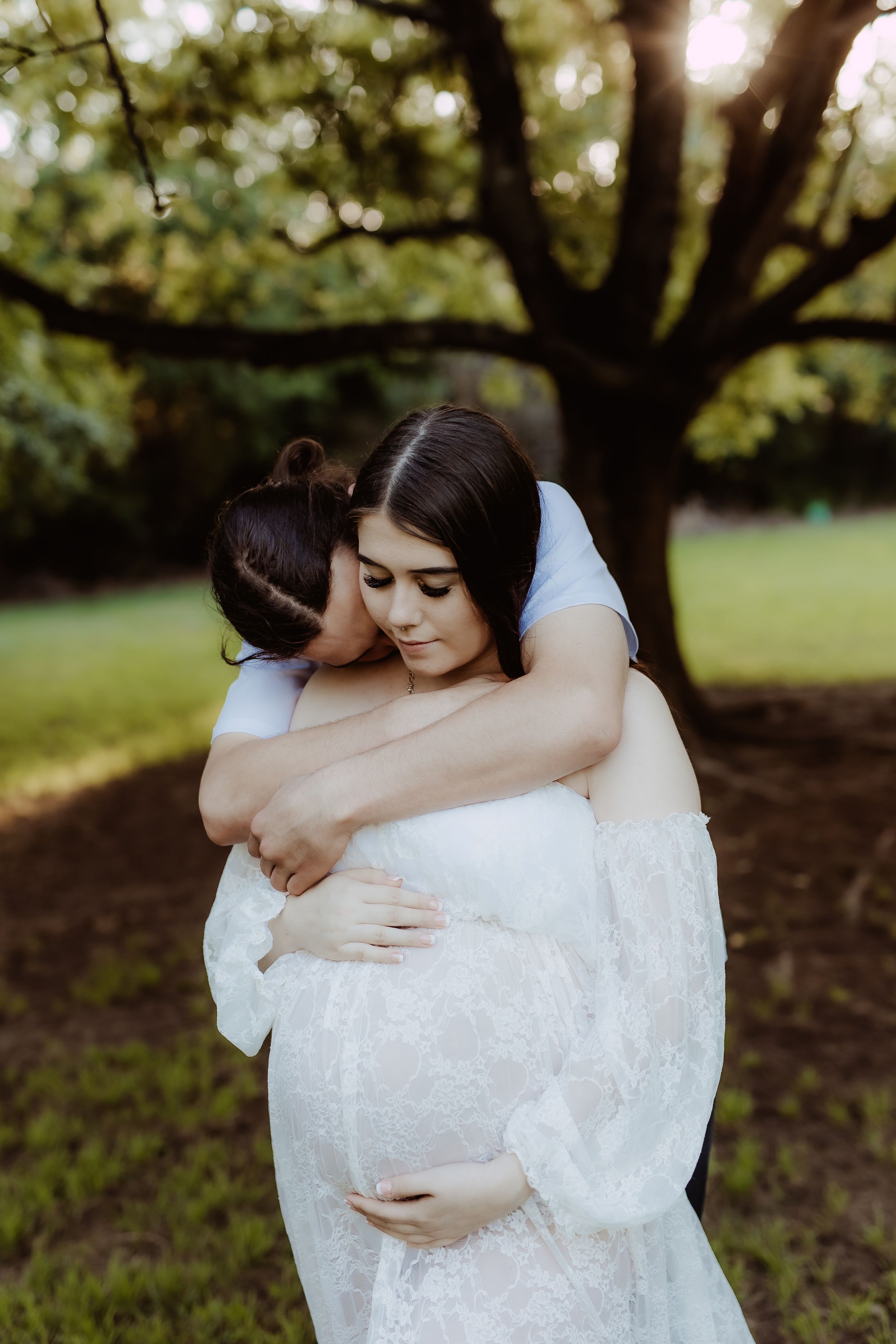 Intimate outdoor maternity session with white lace dress under tree canopy.