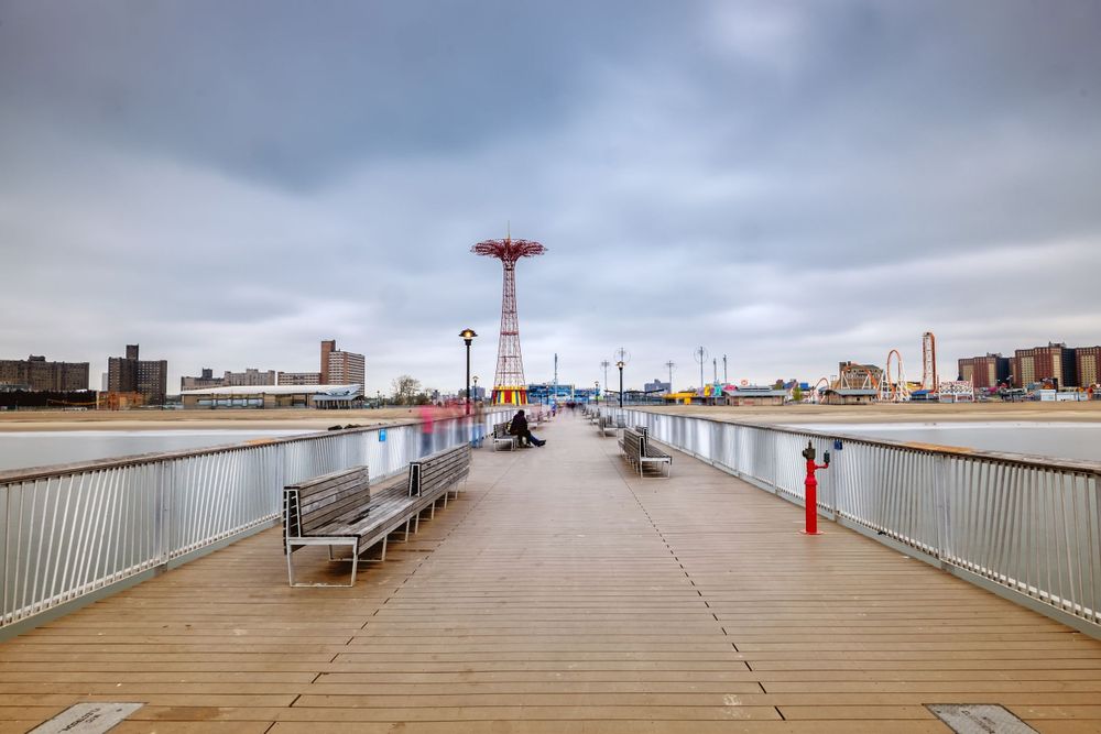 A long boardwalk pier with benches leading to the iconic Coney Island Parachute Jump tower.