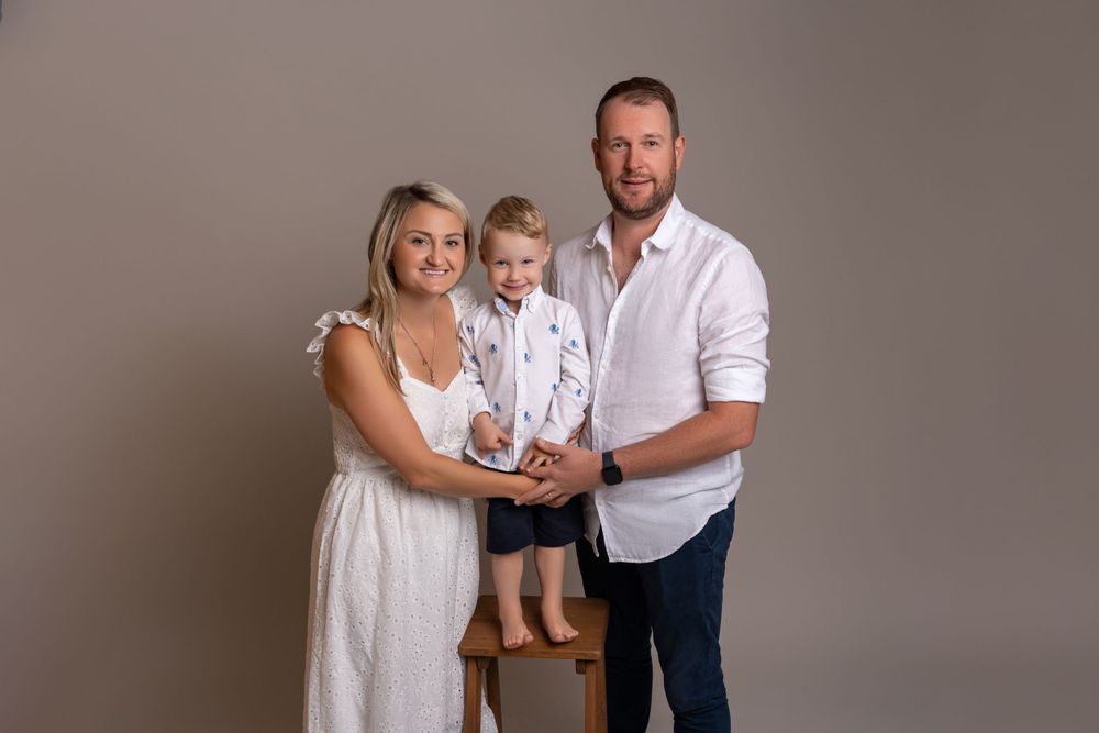 Family of three in white and casual attire posed against gray backdrop.