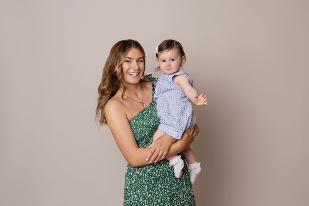 Mother in green patterned dress holds baby in blue outfit against neutral background.