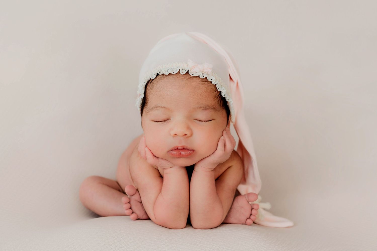 Retrato de una recién nacida en la pose de la ranita con un gorro dormilón rosa durante una sesión de fotos.