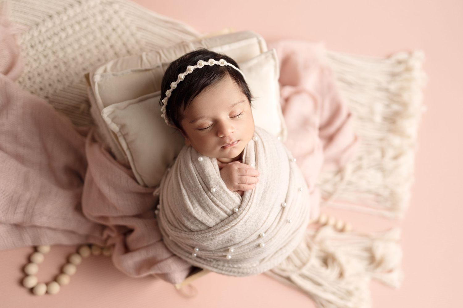 Sleeping newborn wrapped in soft fabric poses on pink textured cushion with pearl accessories.