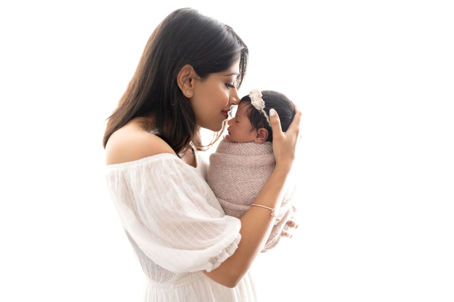 Mother in white dress gently touching noses with swaddled infant in intimate moment.