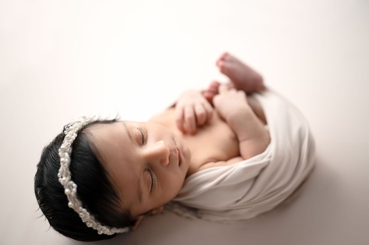 Newborn wearing delicate headband lies peacefully on white backdrop during portrait session.