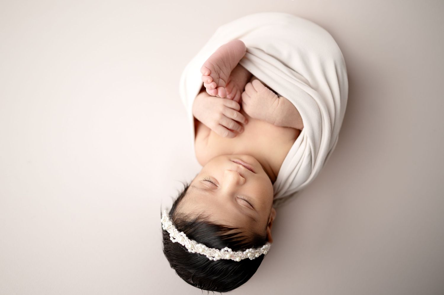 Sleeping newborn wrapped in white fabric lies against neutral background during professional photo session.