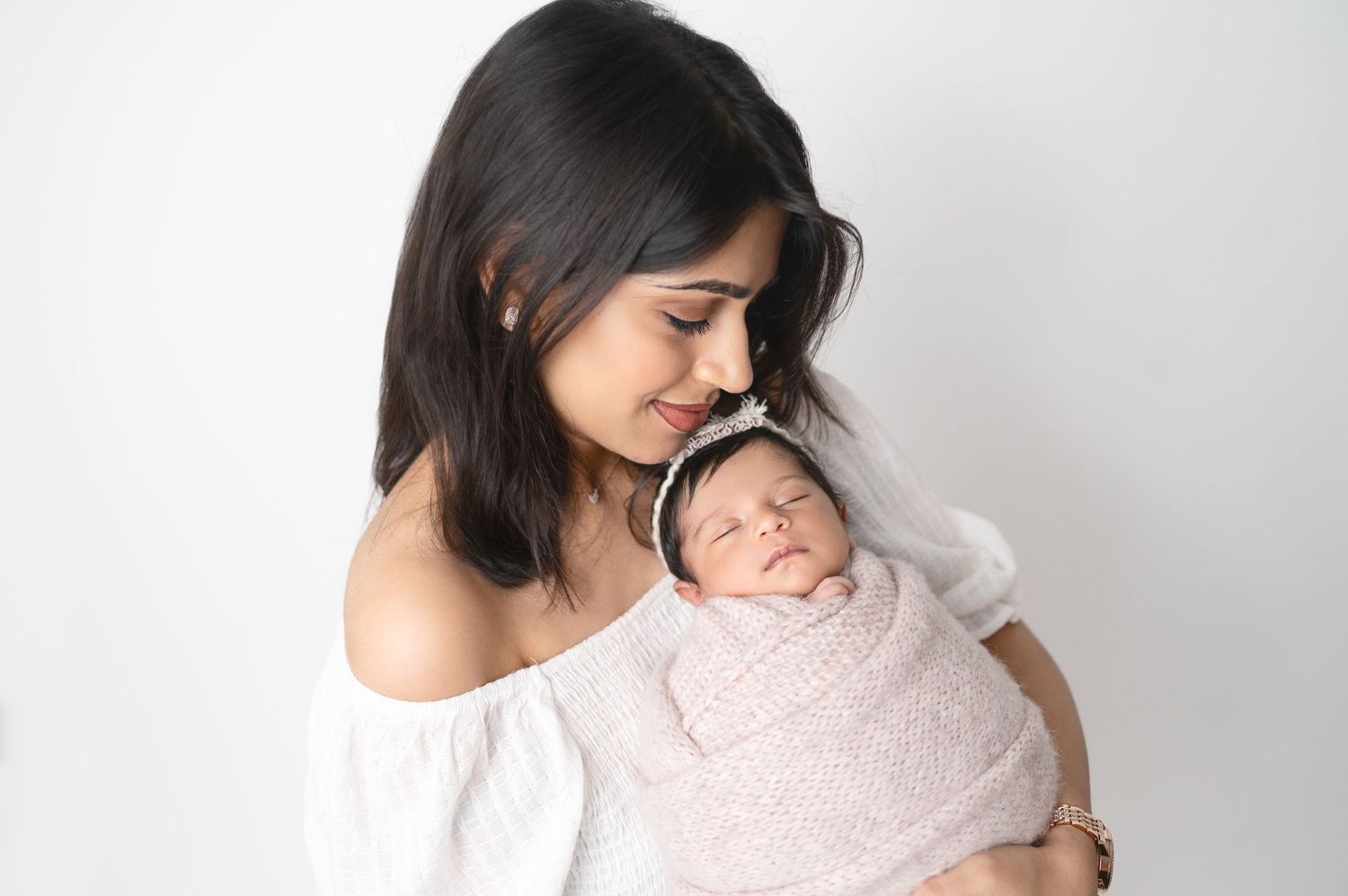 Mother in white top holds swaddled newborn tenderly against simple backdrop.