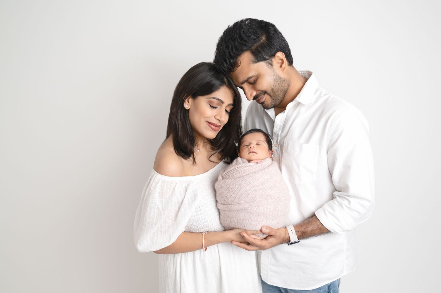 Parents in white outfits tenderly cradling their swaddled newborn against white background.