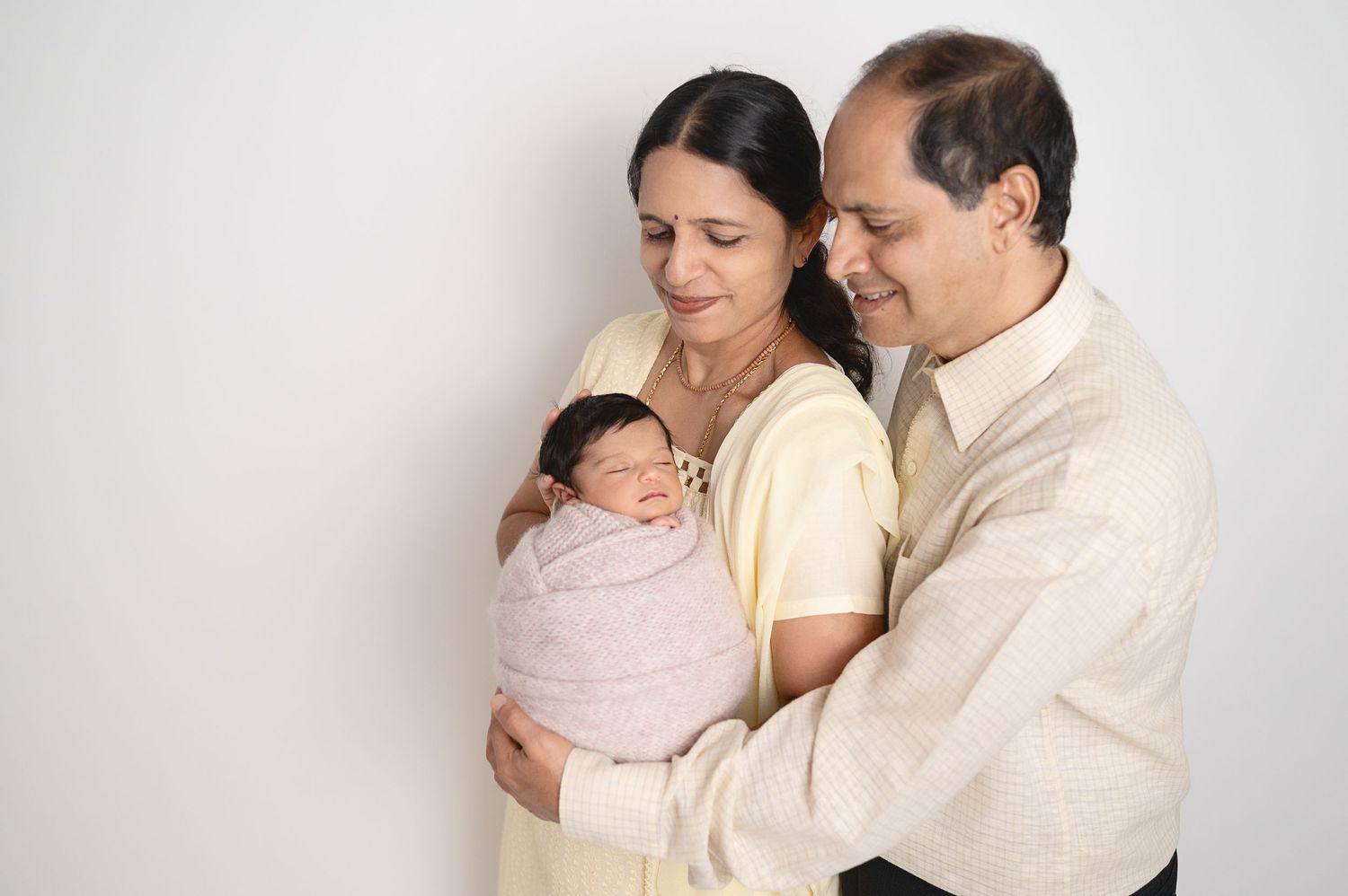 Parents lovingly embrace their swaddled newborn baby against white studio backdrop.