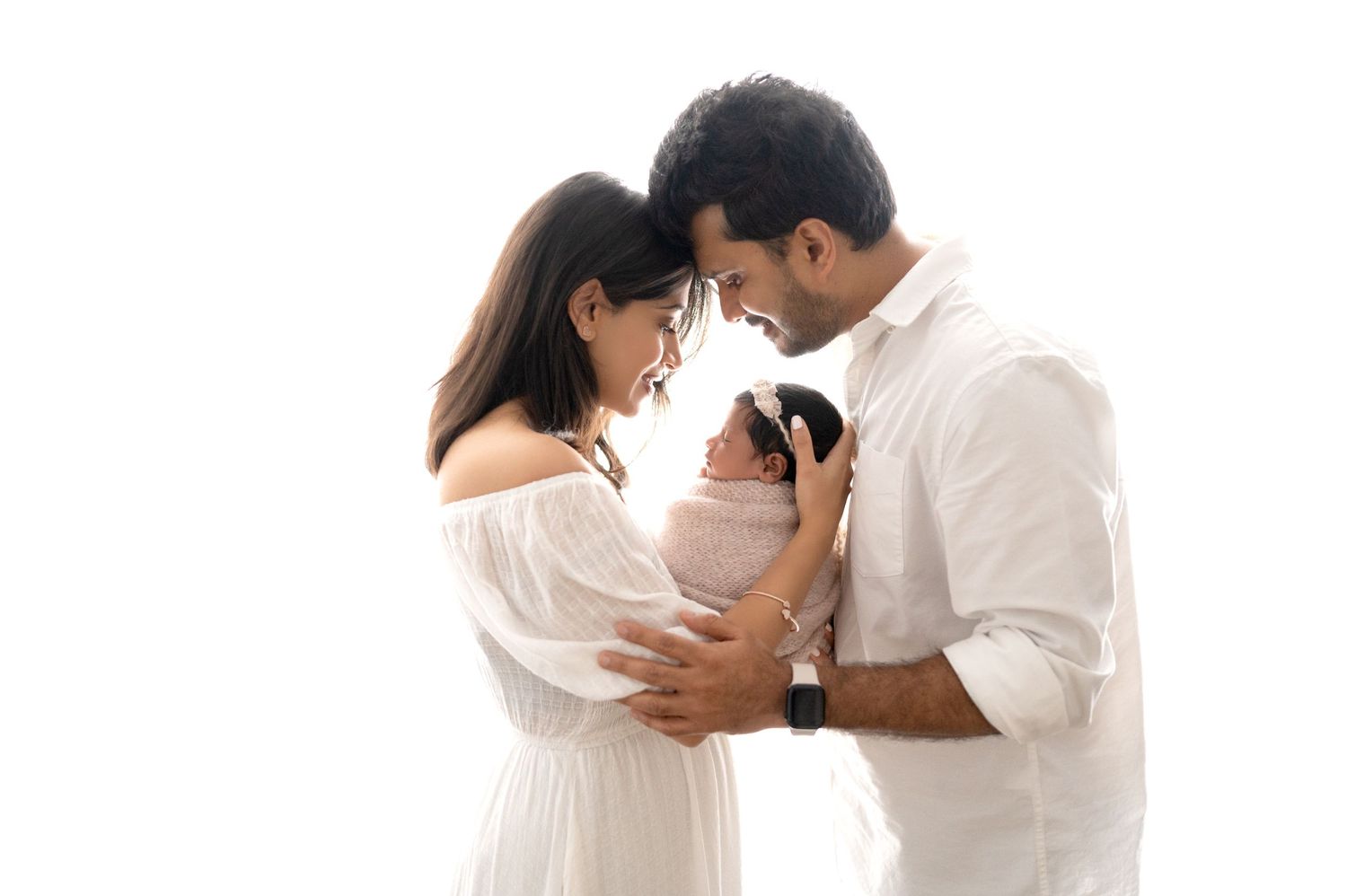 Parents sharing intimate moment with newborn against white backdrop in matching white outfits.