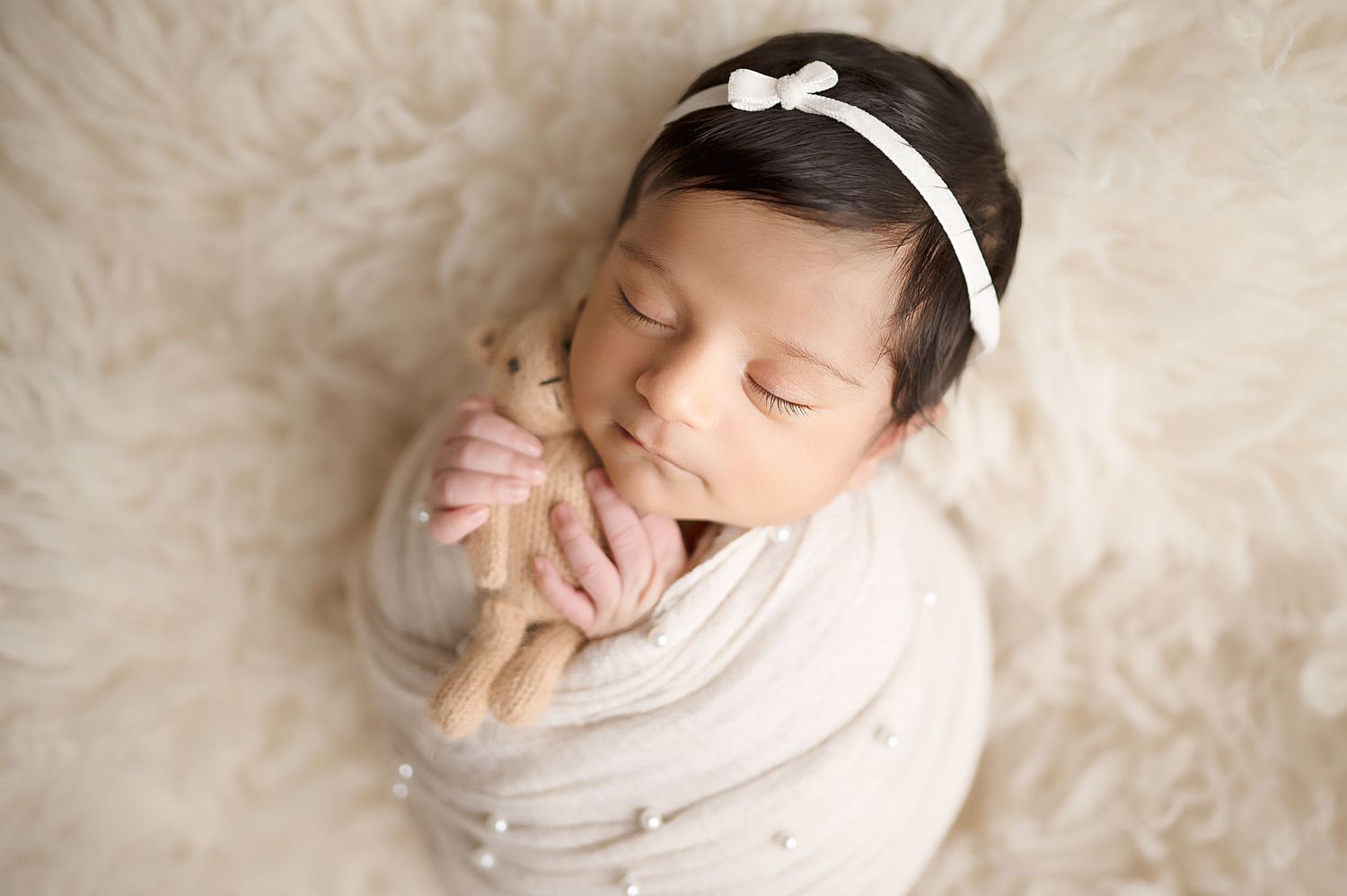 Peaceful newborn baby in white wrap and headband rests on soft furry backdrop.