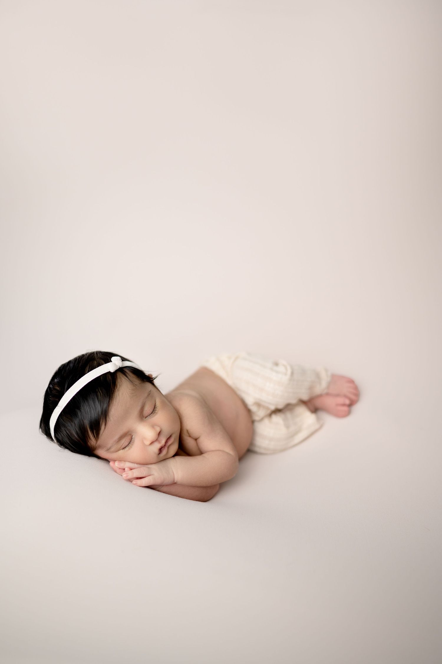 Peaceful newborn sleeping on side wearing white headband against minimal beige backdrop.