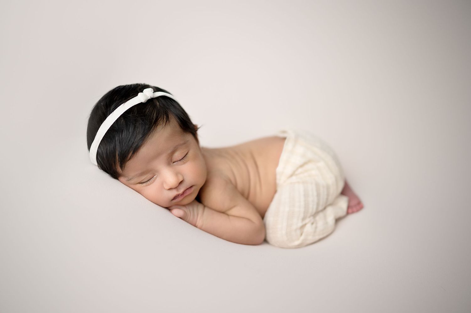 Sleeping newborn with white headband rests peacefully in cream-colored wrap.