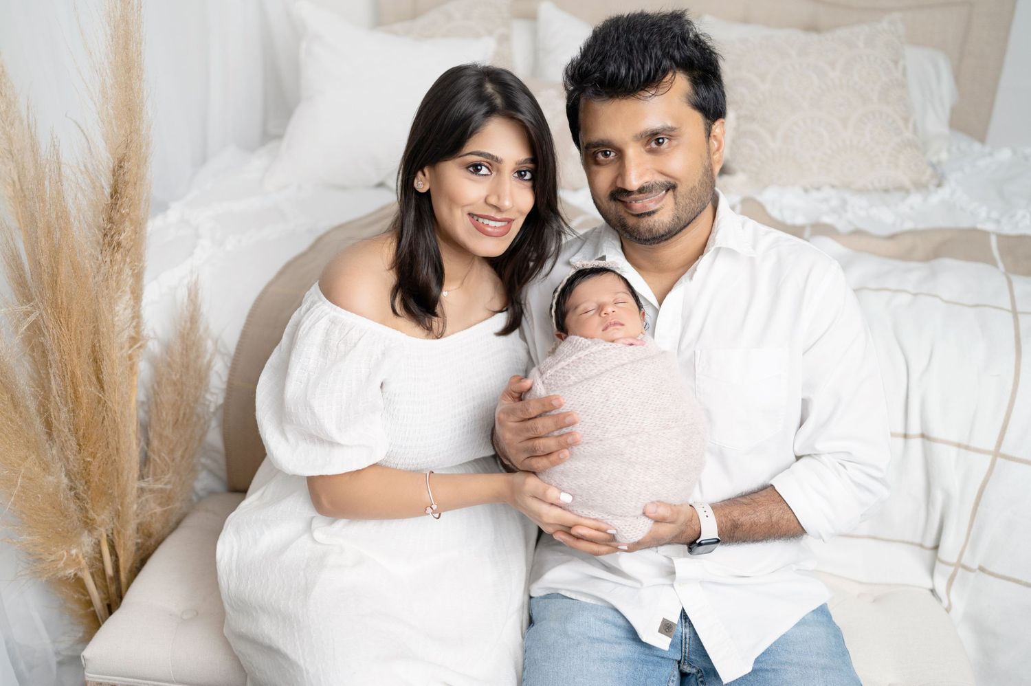 A couple in white attire poses with their newborn baby against a light backdrop with decorative pampas grass.