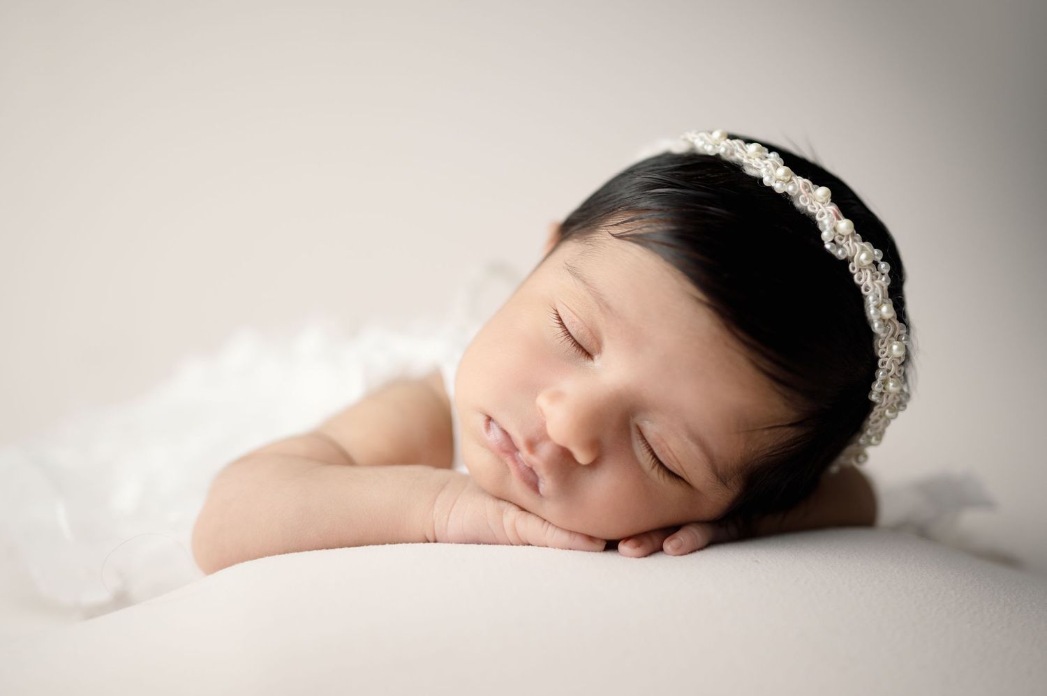Sleeping baby with ornate headband rests on white blanket during newborn photography session.