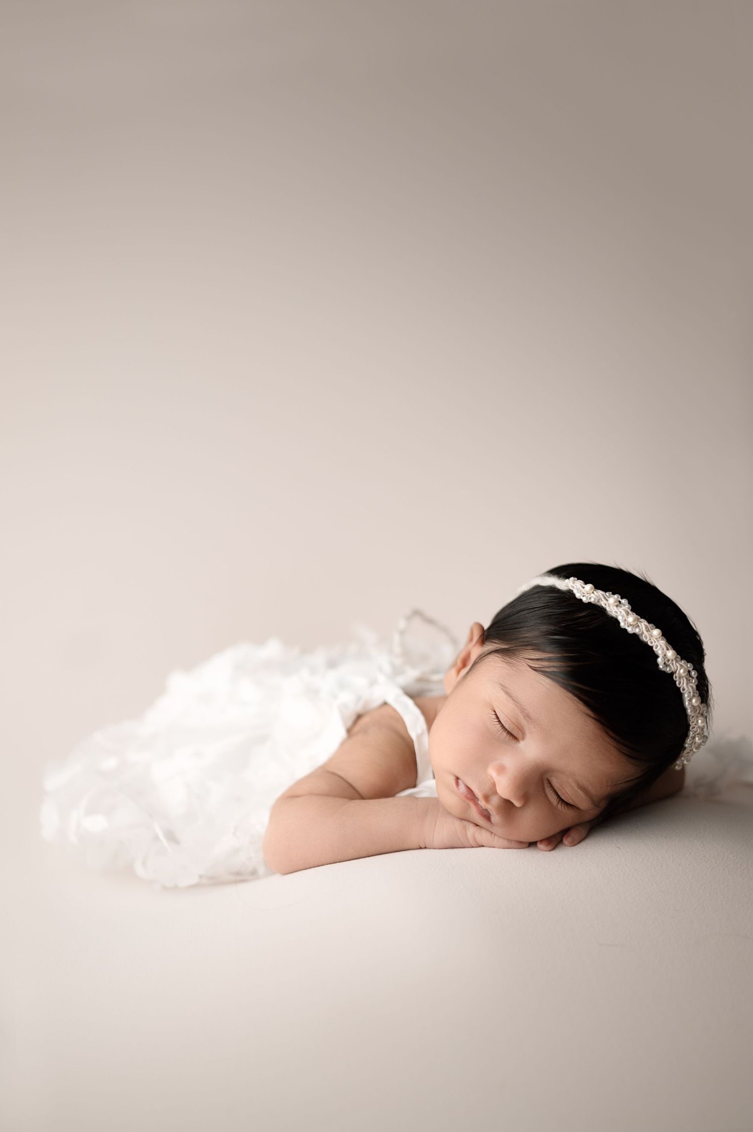Peaceful newborn in white dress and headband sleeps during professional portrait session.