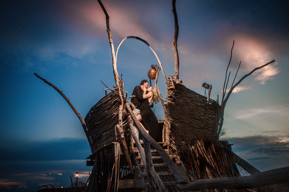 Artistic wooden structure on beach at sunset with dramatic sky and driftwood architecture.