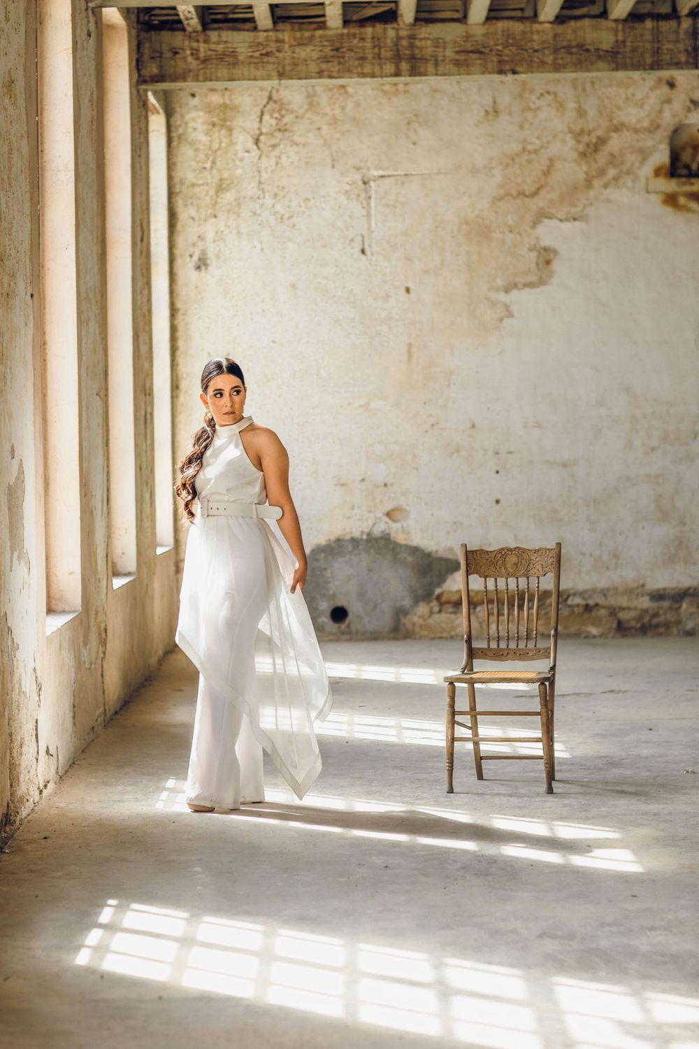 A graceful figure in a white gown poses elegantly in a sunlit corridor with a vintage wooden chair.