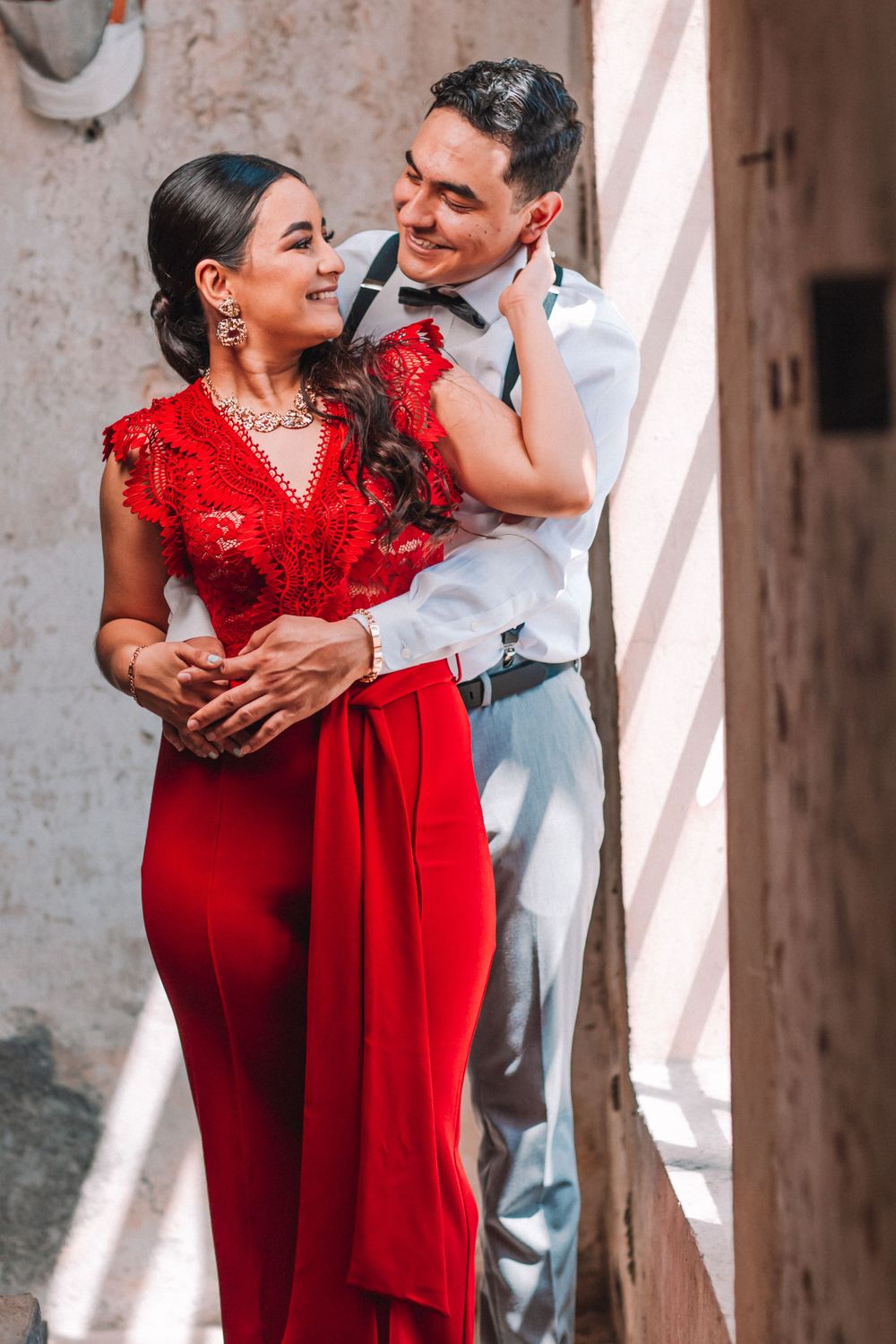 A romantic couple embraces against a rustic wall, with the woman wearing a stunning red lace dress.