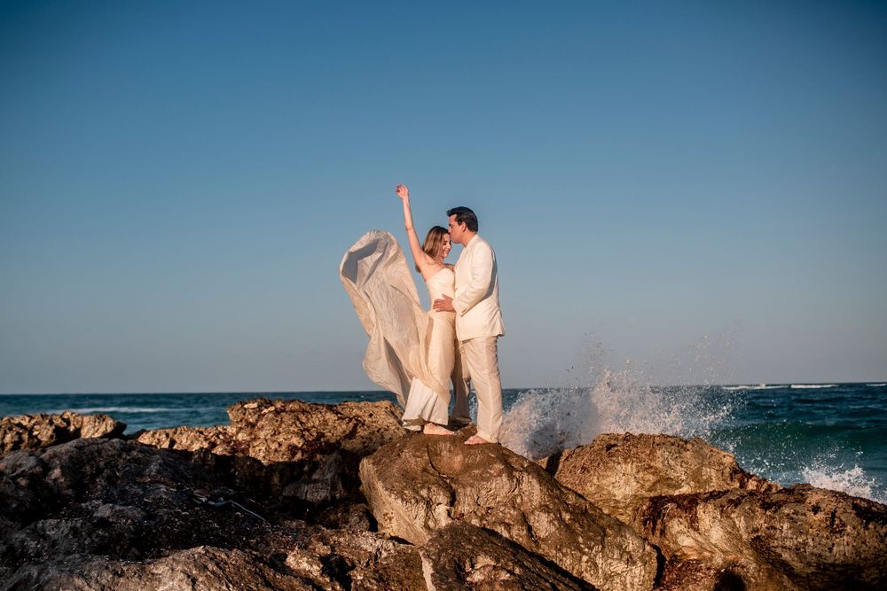 A couple in light-colored attire embraces on rocky coastal cliffs with crashing waves in the background.