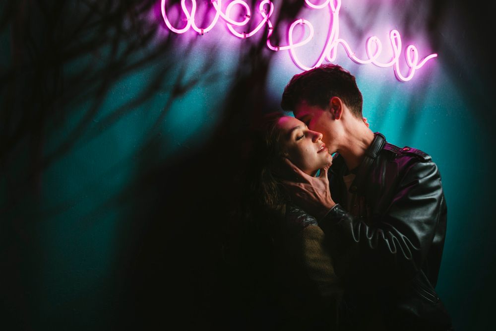 A romantic couple shares an intimate moment under a pink neon sign in moody lighting.