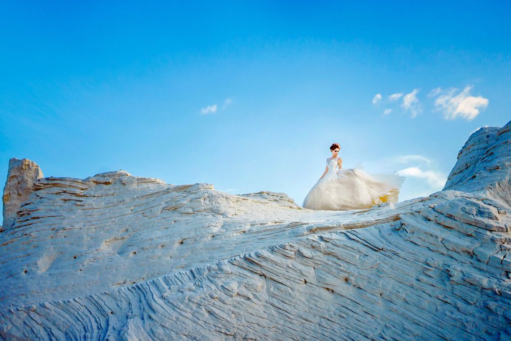 White gown flowing dramatically on sandy white cliff against blue sky.