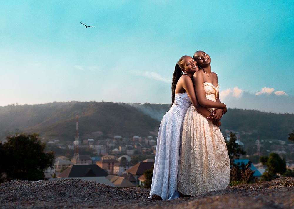Romantic couple in white dresses embracing on hilltop with cityscape and birds in background.