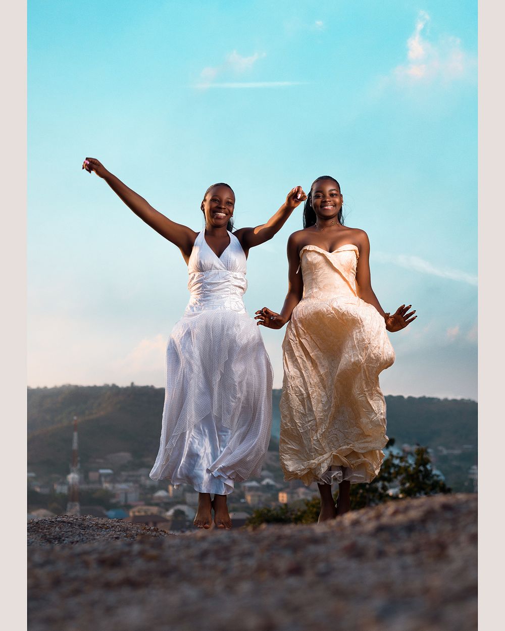 Two people in flowing dresses jumping joyfully against blue sky and mountain landscape.