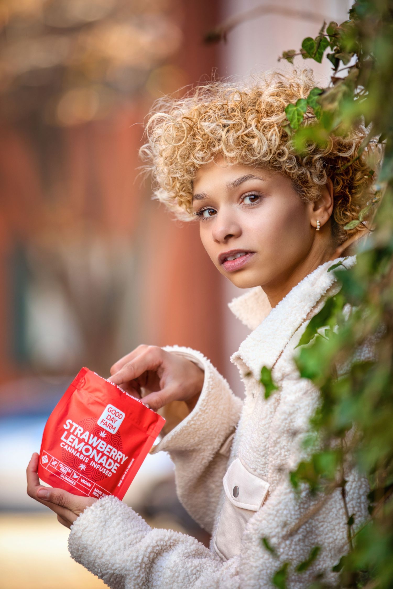 Young woman with curly blonde hair holding red Strawberry Lemonade cannabis package outdoors.
