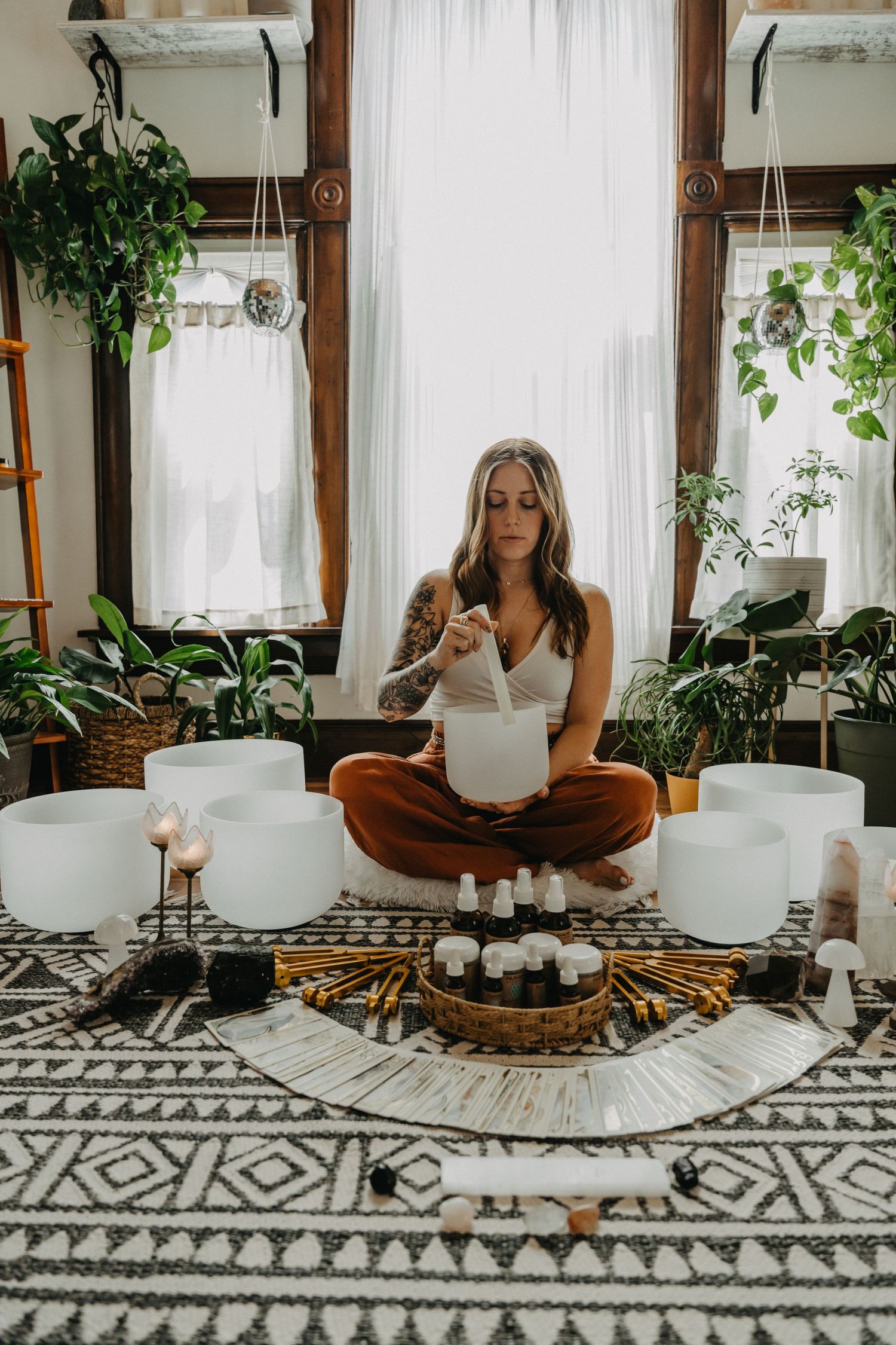 Woman in rust pants playing crystal singing bowls surrounded by candles and plants in a boho room.