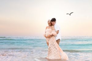 Romantic couple in flowy outfits embraces on beach as seagull flies overhead at sunset.