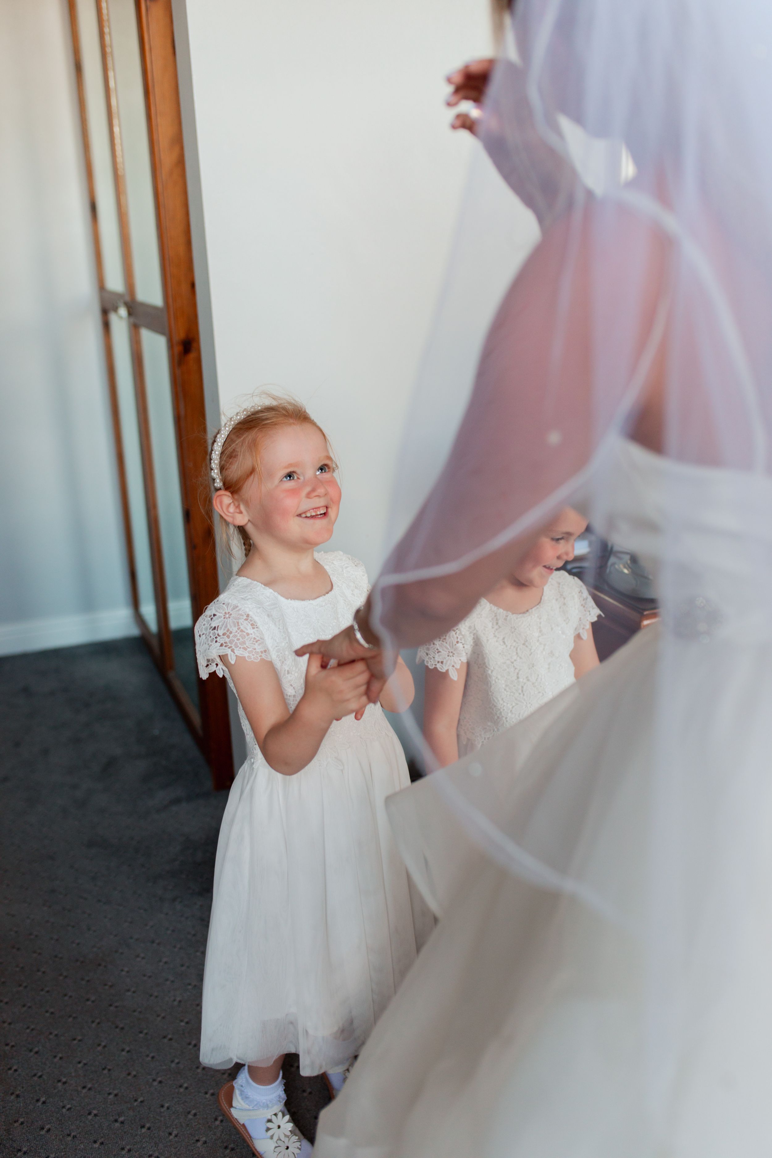 A young flower girl in white lace dress smiles while looking up at a wedding gown. Waterton Park Hotel.