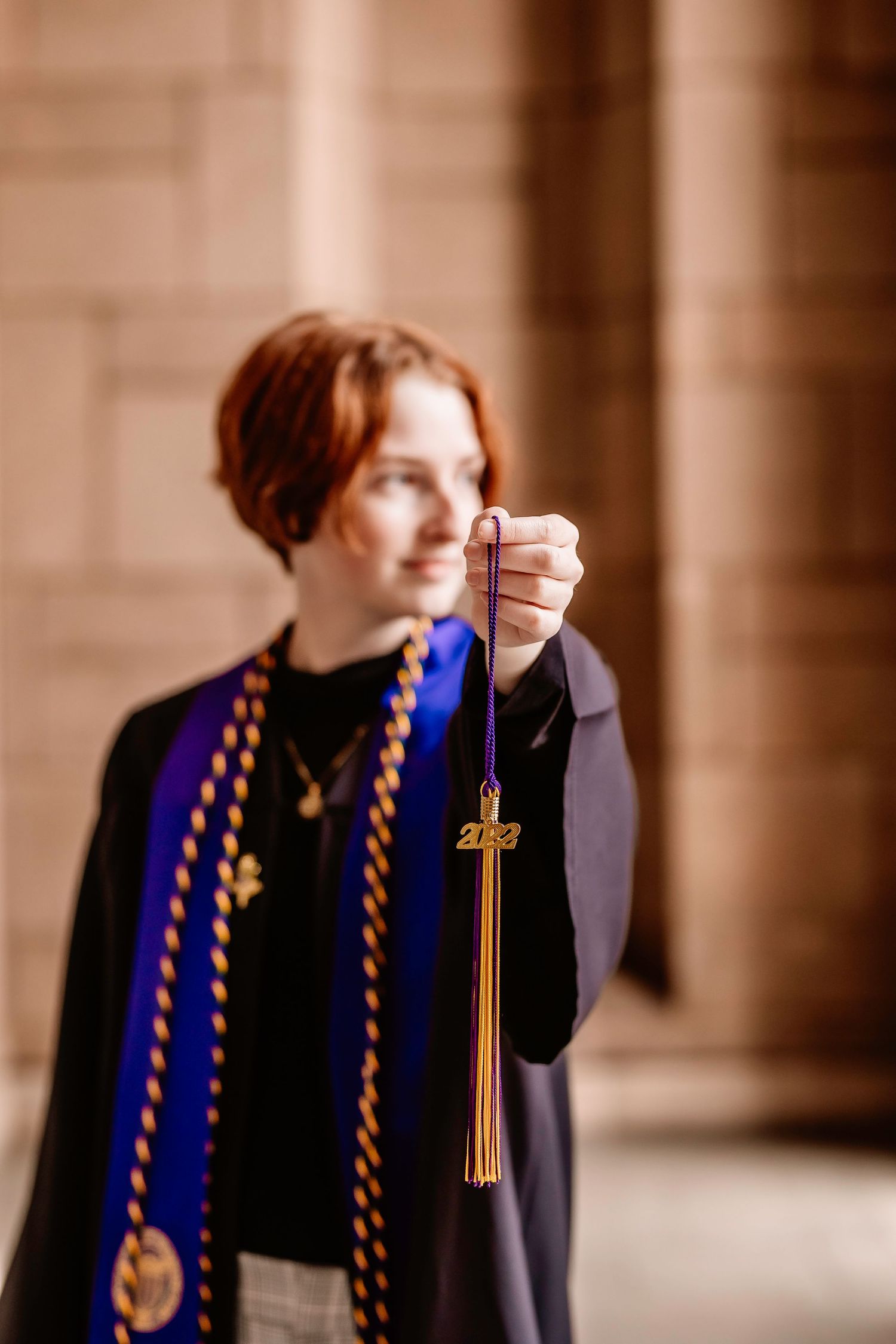 Academic wearing blue stole holds up graduation tassel charm in stone hallway.