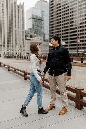 Stylish couple in casual wear walking together on urban boardwalk with skyscrapers in background.