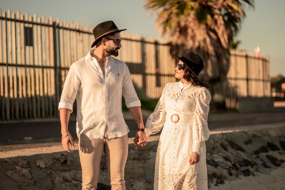 Couple in coordinated white and beige outfits stroll along beachfront with palm trees.