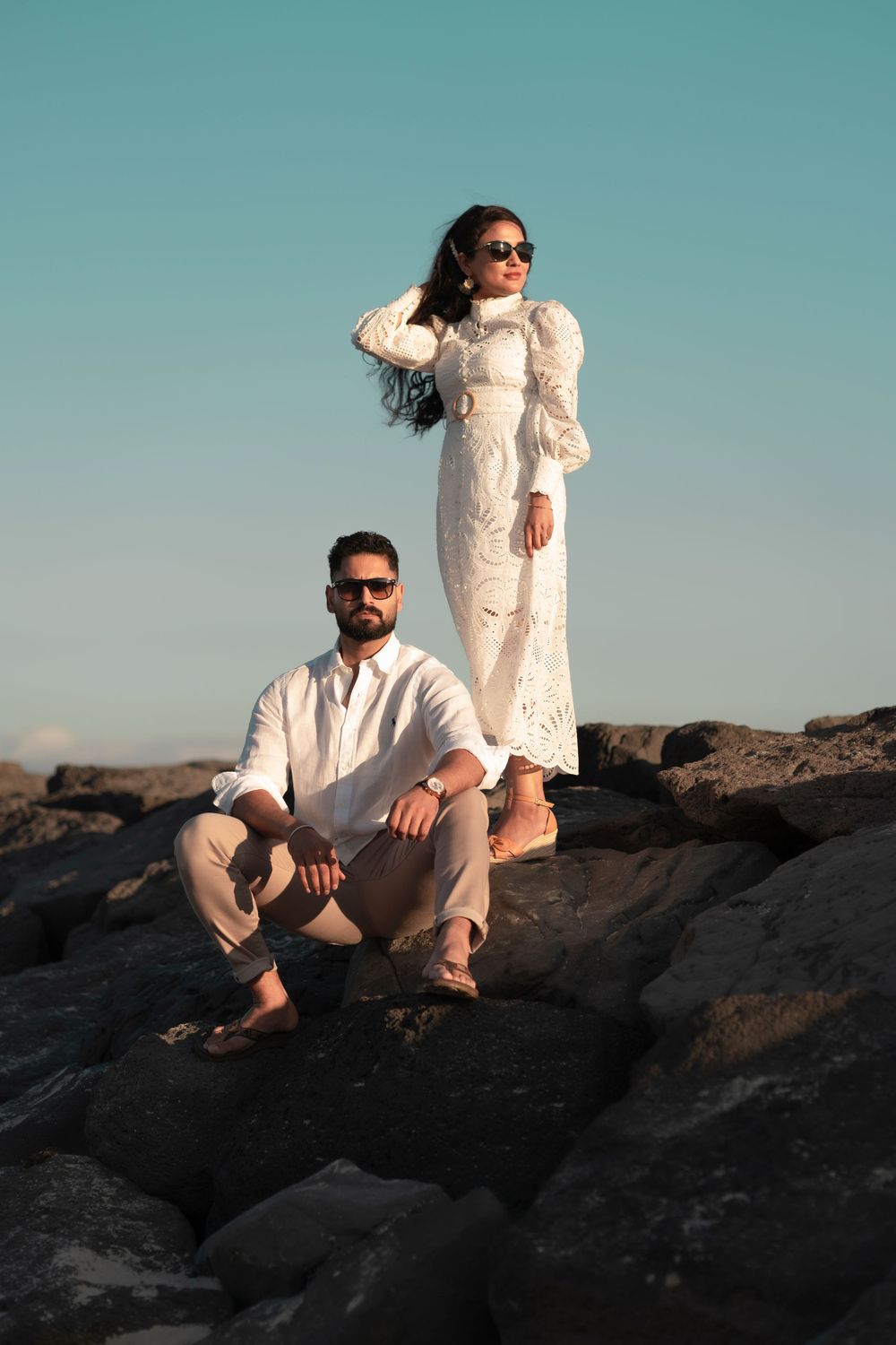Stylish couple in white outfits pose dramatically on rocky coastal formation against blue sky.