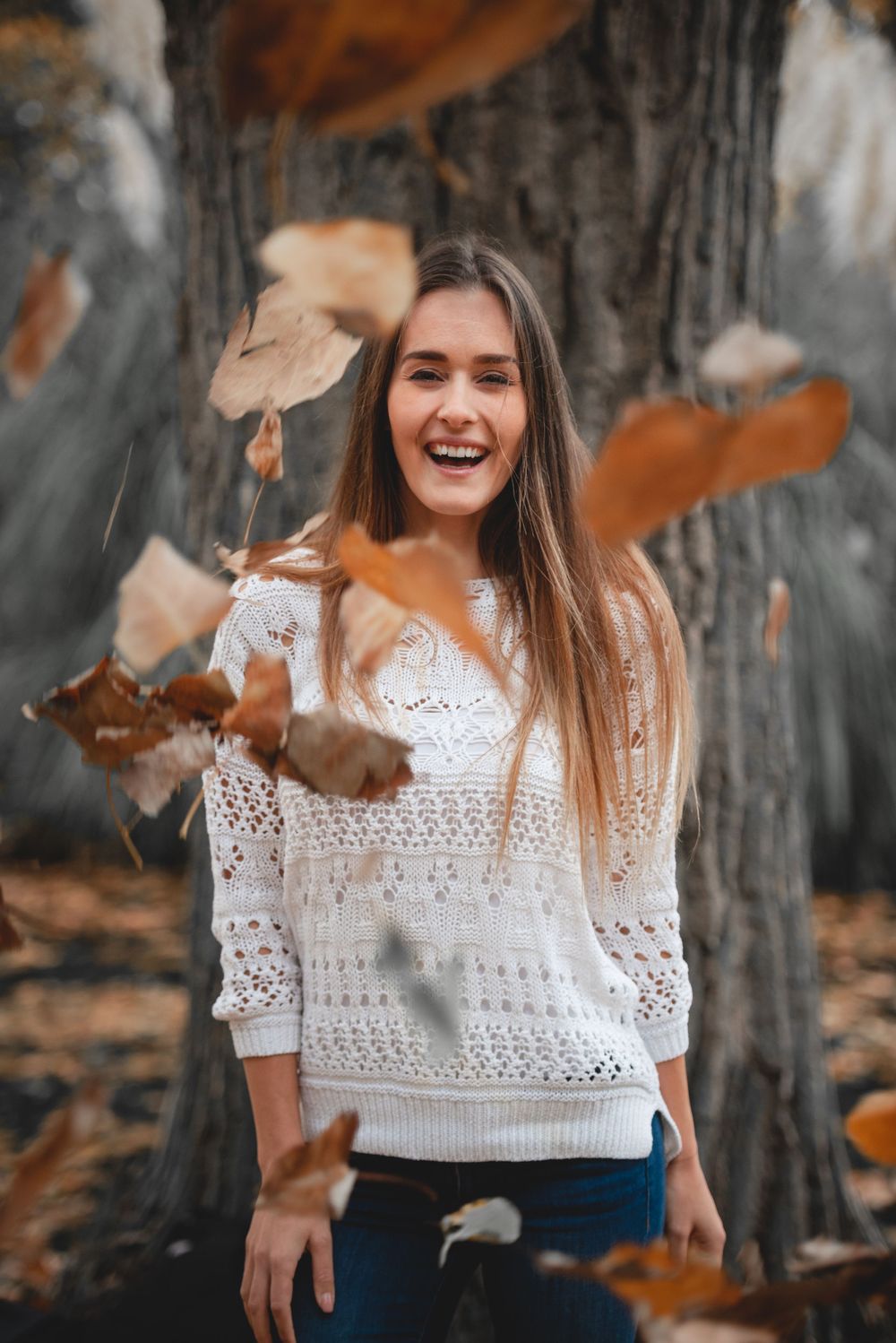 A person in a white crochet sweater laughs joyfully as autumn leaves fall around them in a forest setting.