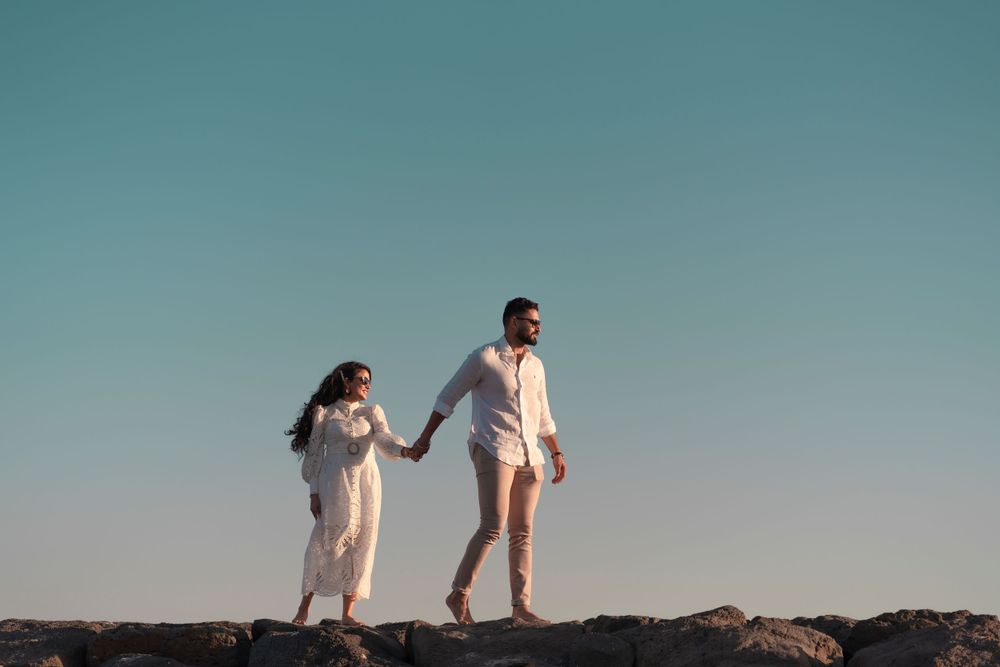 Couple in white clothing walks along rocky coastal path during sunset photoshoot.