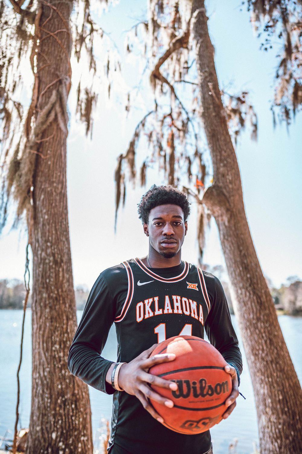 Basketball player in Oklahoma jersey holds Wilson ball among Spanish moss trees.