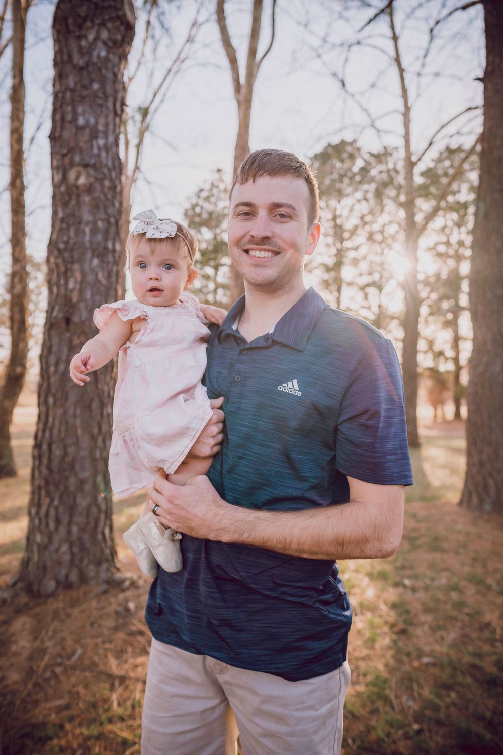 Father holding smiling baby girl in pink dress while standing in wooded area.