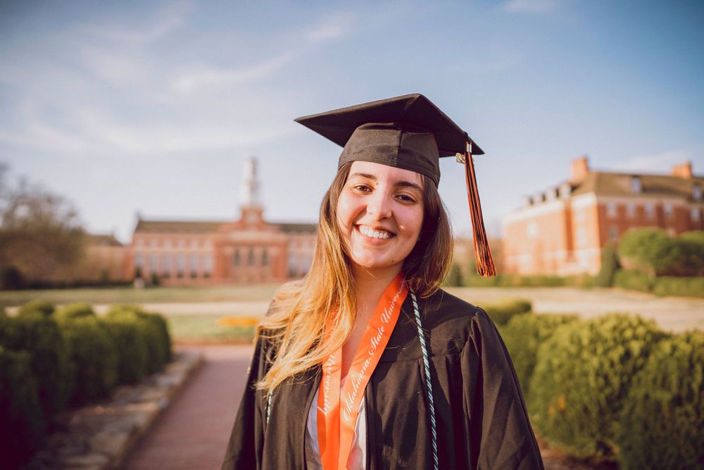 Graduate in traditional cap and gown smiles brightly on campus with brick buildings in background.