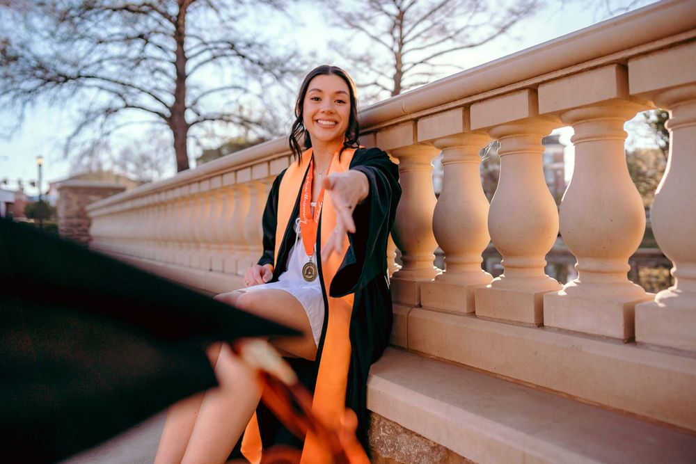 Graduate in academic dress sits on ornate stone railing during golden hour photo session.