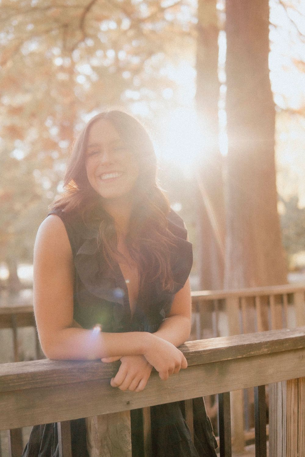 Woman in black dress laughing while leaning on wooden railing with golden sunlight filtering through trees.
