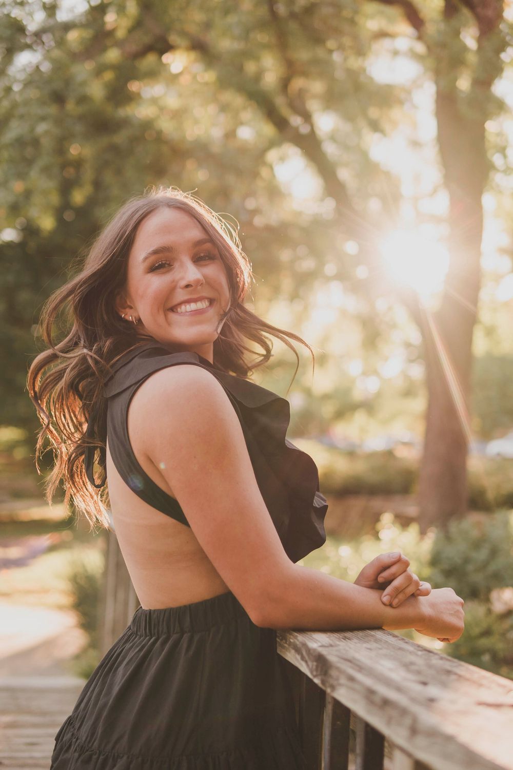 A golden hour portrait showing someone in a black outfit smiling over their shoulder on a wooden bridge.