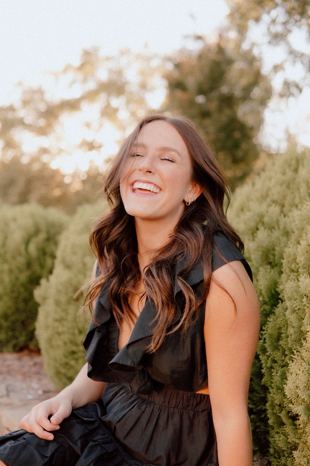 Woman in black dress laughing joyfully against background of green shrubs.